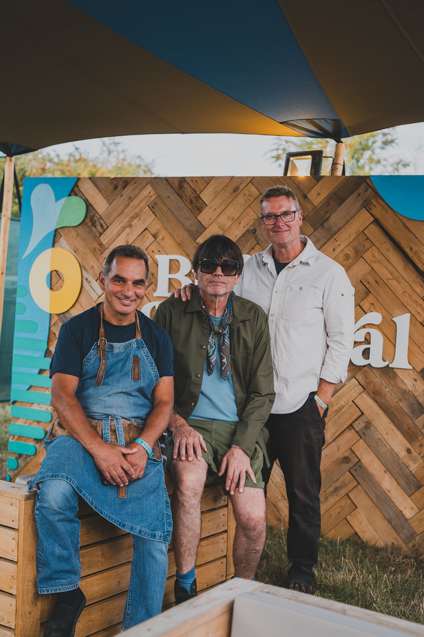 Three men posing together in front of a rustic wooden herringbone backdrop at a food festival.