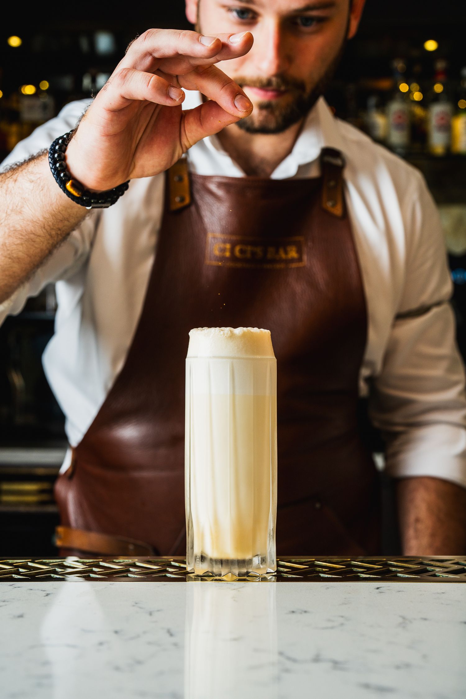 Bartender garnishing a creamy white frothy cocktail in tall glass at upscale bar.