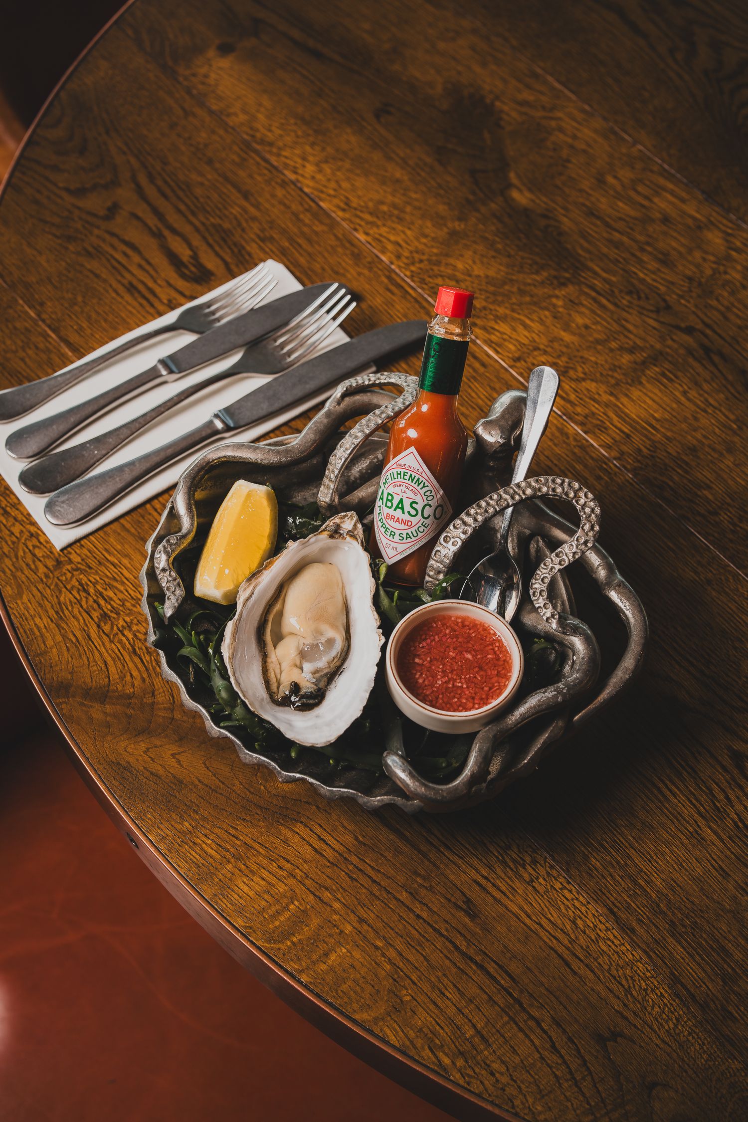 Single oyster served in decorative metal dish with Tabasco, dipping sauce, and lemon on wood table.