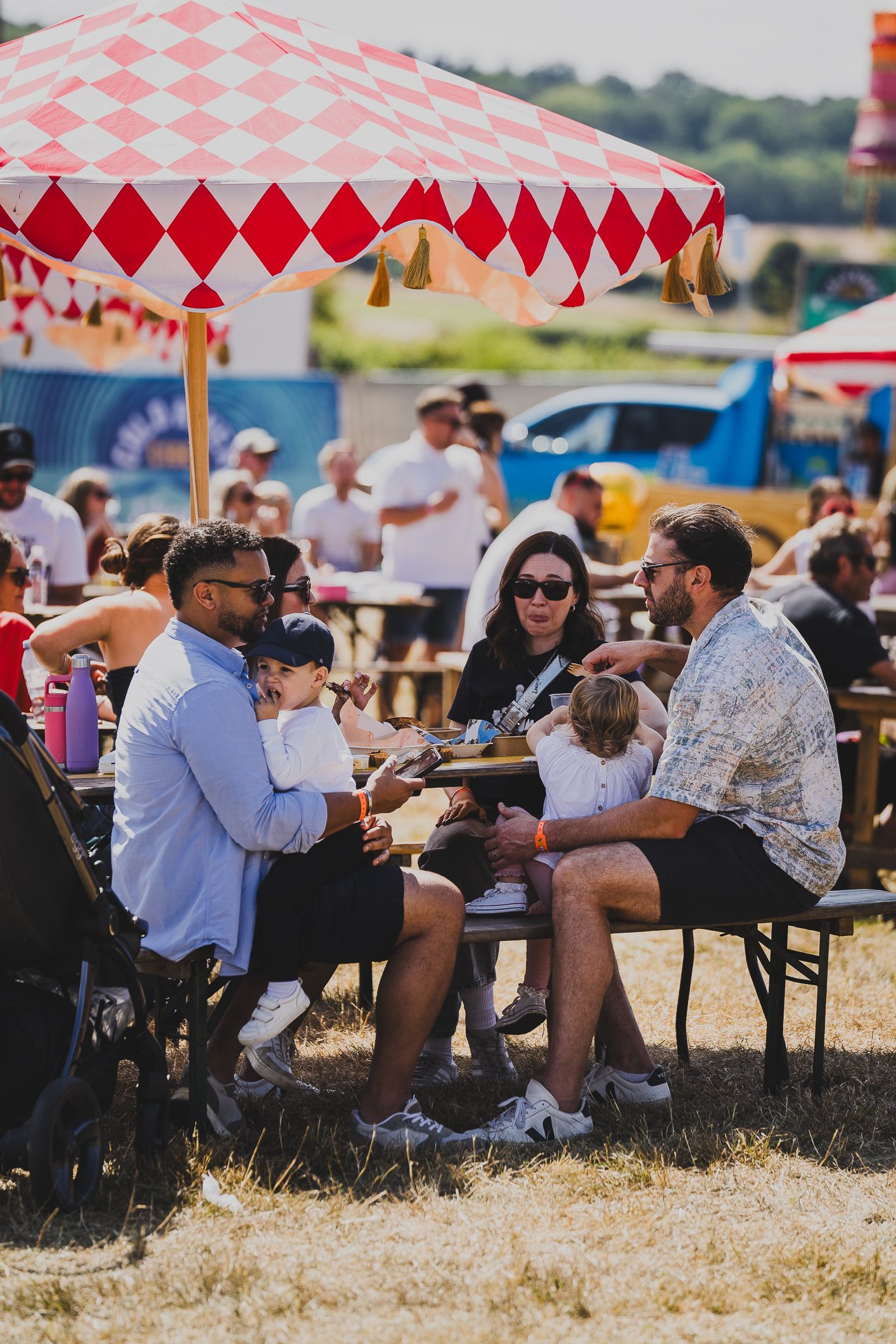 Families dining at outdoor tables under red and white striped umbrellas at a festival.