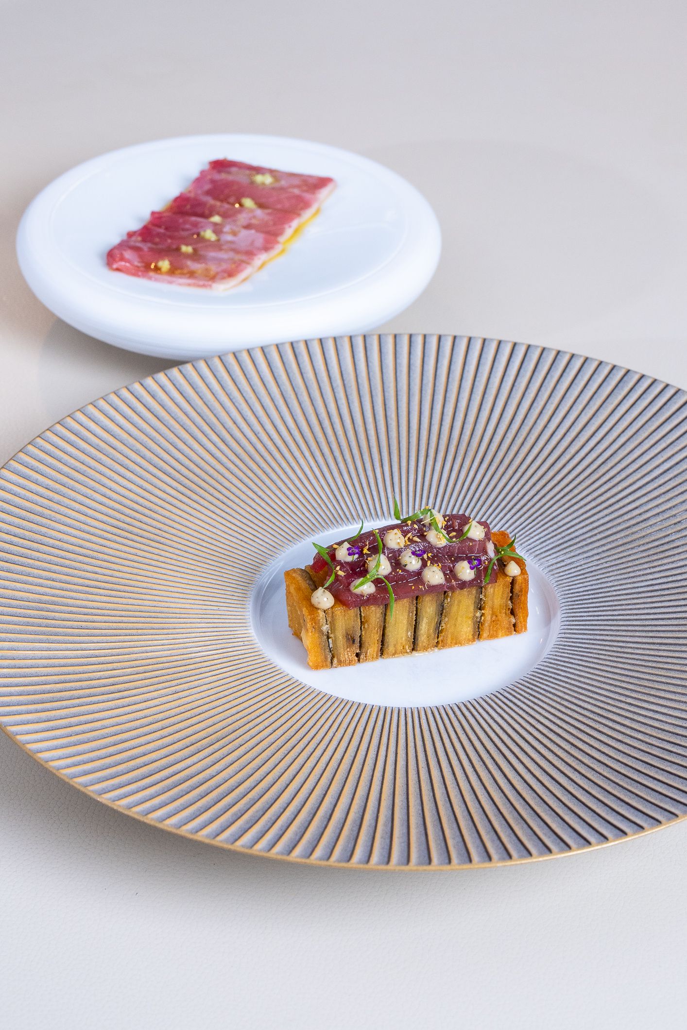 Elegant tuna tartare on striped gold-rimmed plate with a second serving blurred in background.