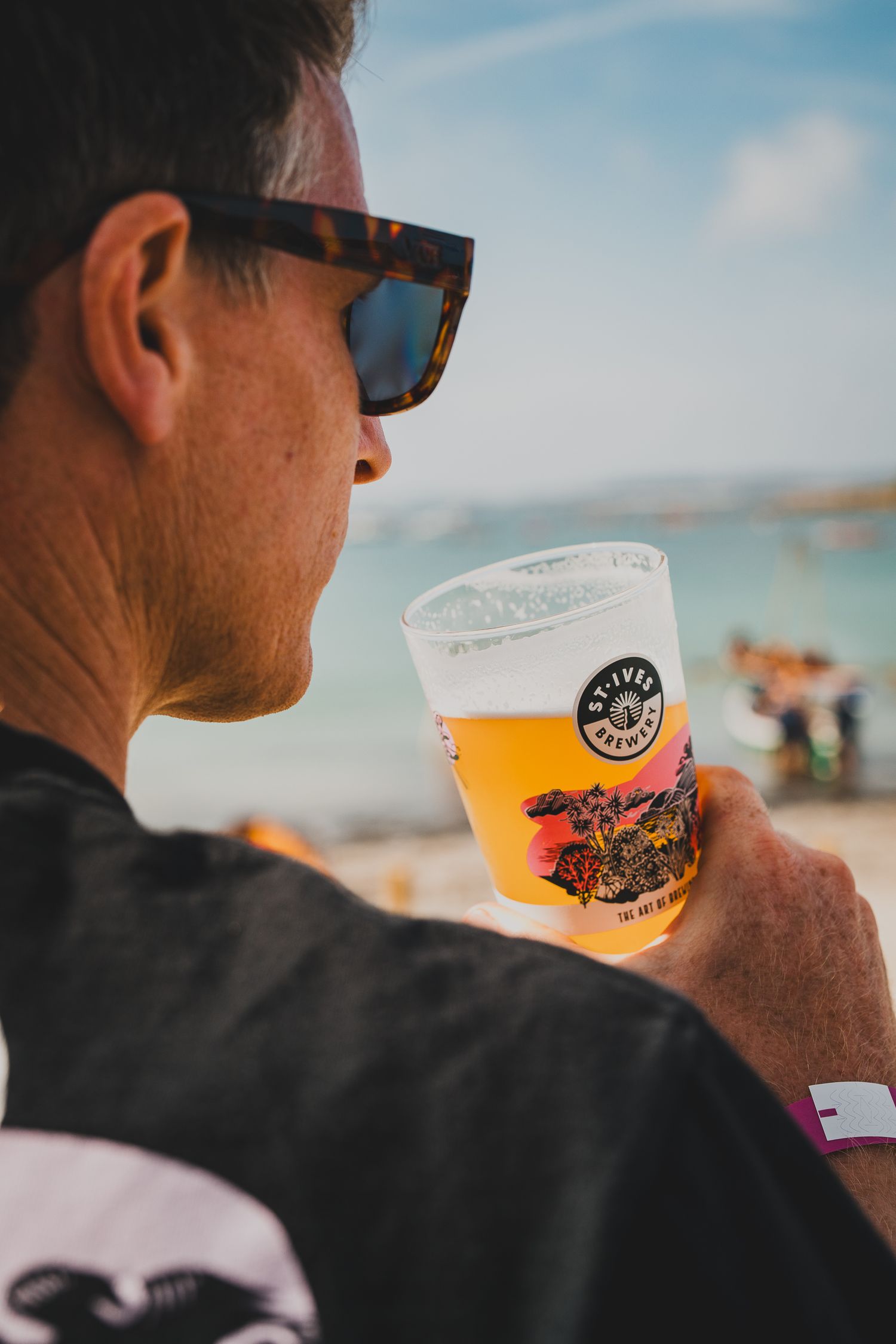 Man in sunglasses holding a craft beer cup at a sunny beach festival.
