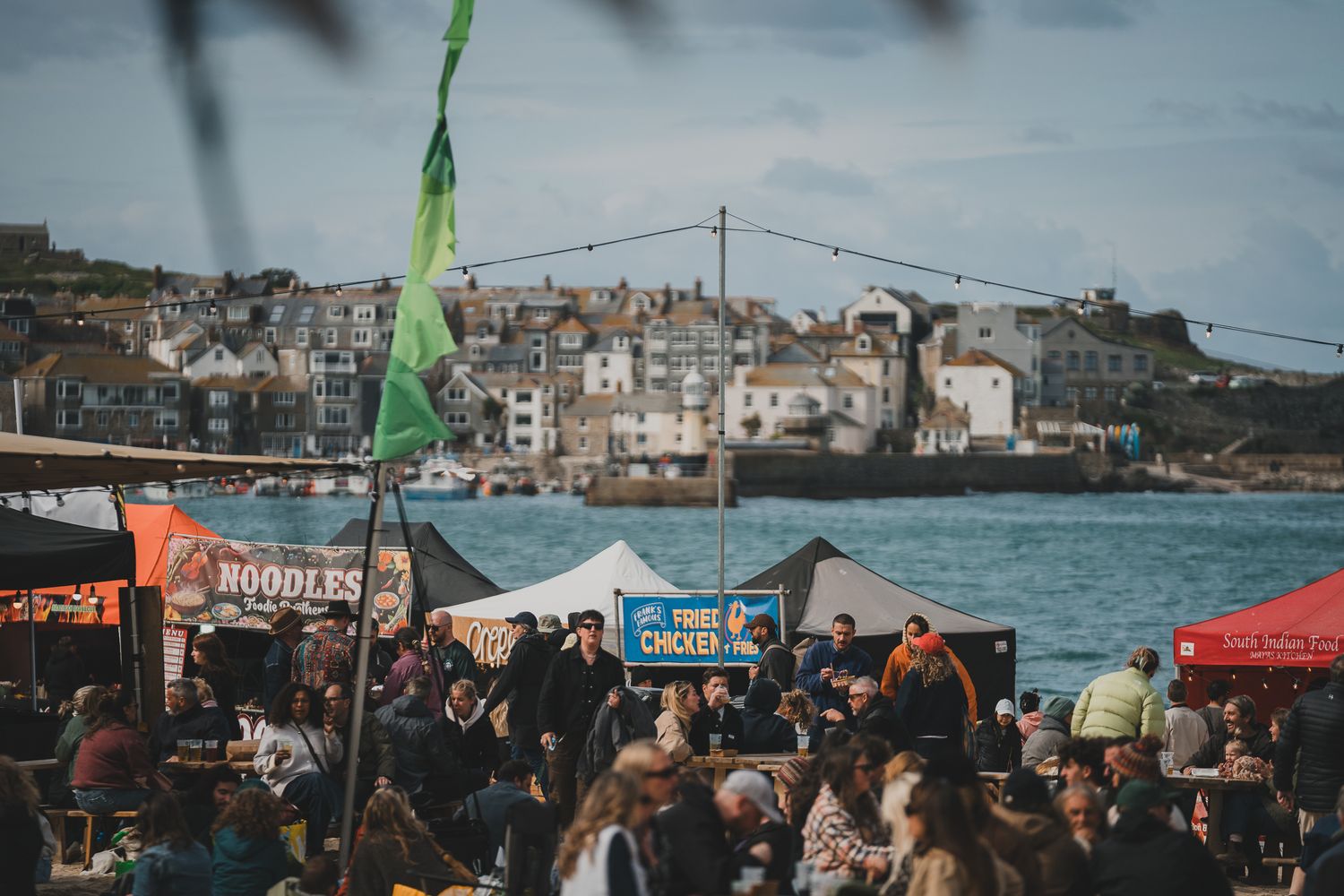 Busy waterfront food festival with noodle and chicken stalls, green flags, and historic town backdrop.