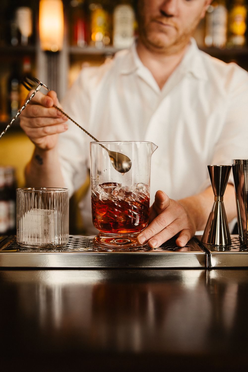 Bartender stirring a dark cocktail with ice in a mixing glass at a bar.