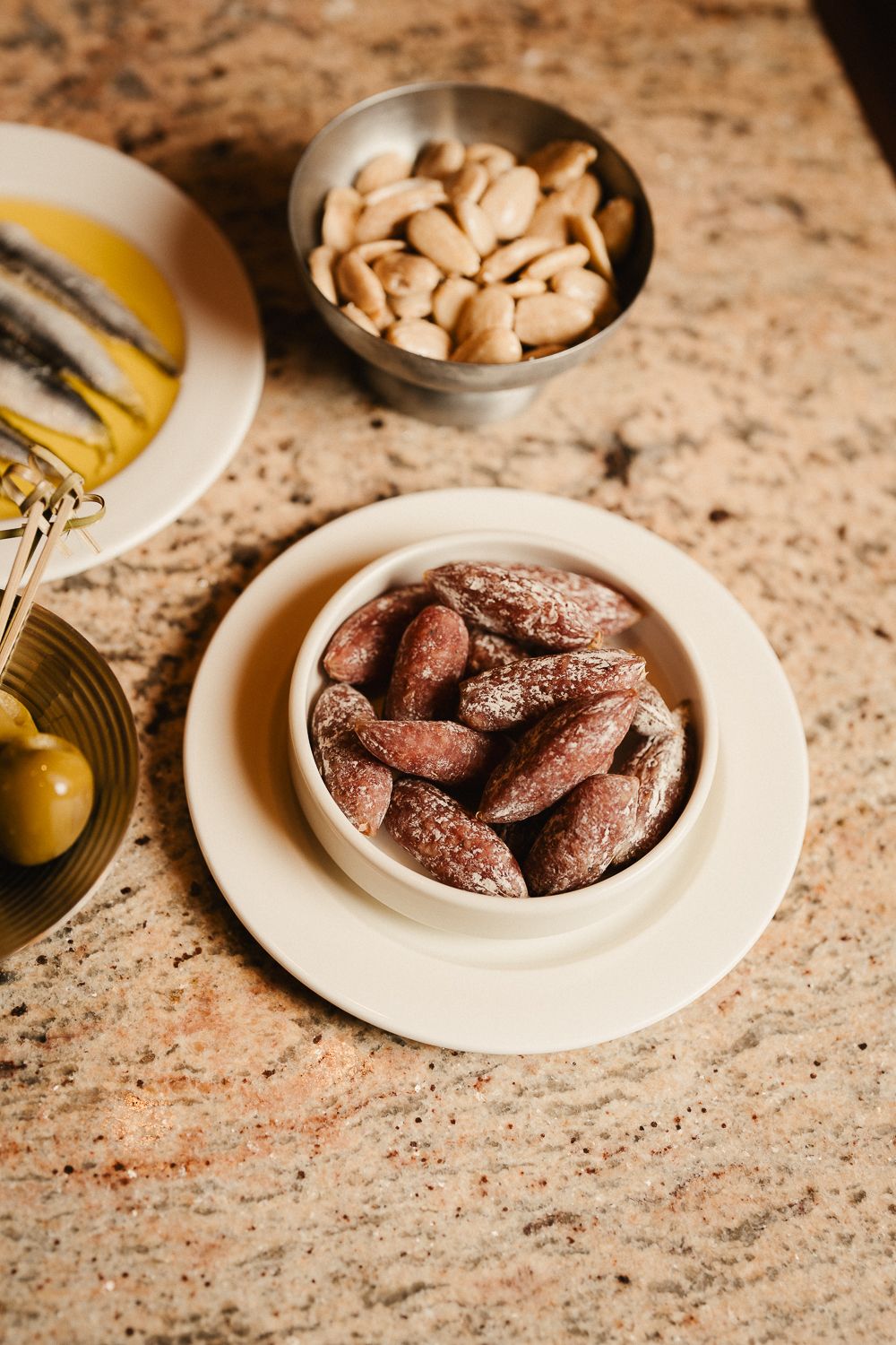 Spiced almonds in a bowl with anchovies and blanched almonds on marble counter.