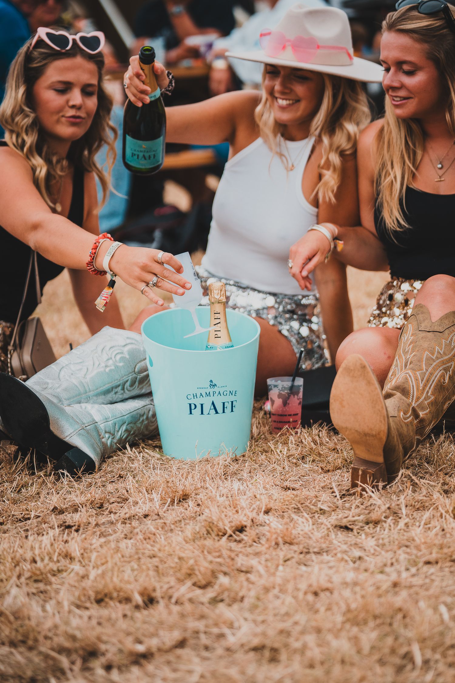 Three women enjoy Champagne Piaff at an outdoor festival, placing bottles in a teal ice bucket on hay.