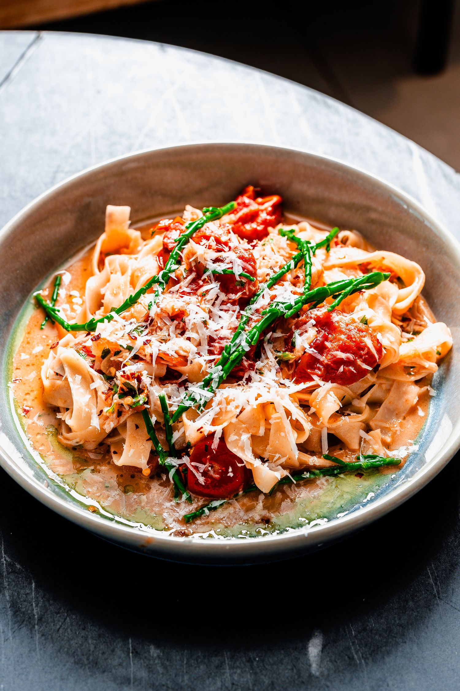 Bowl of fettuccine pasta with tomato sauce, samphire, and grated parmesan cheese on a marble surface.