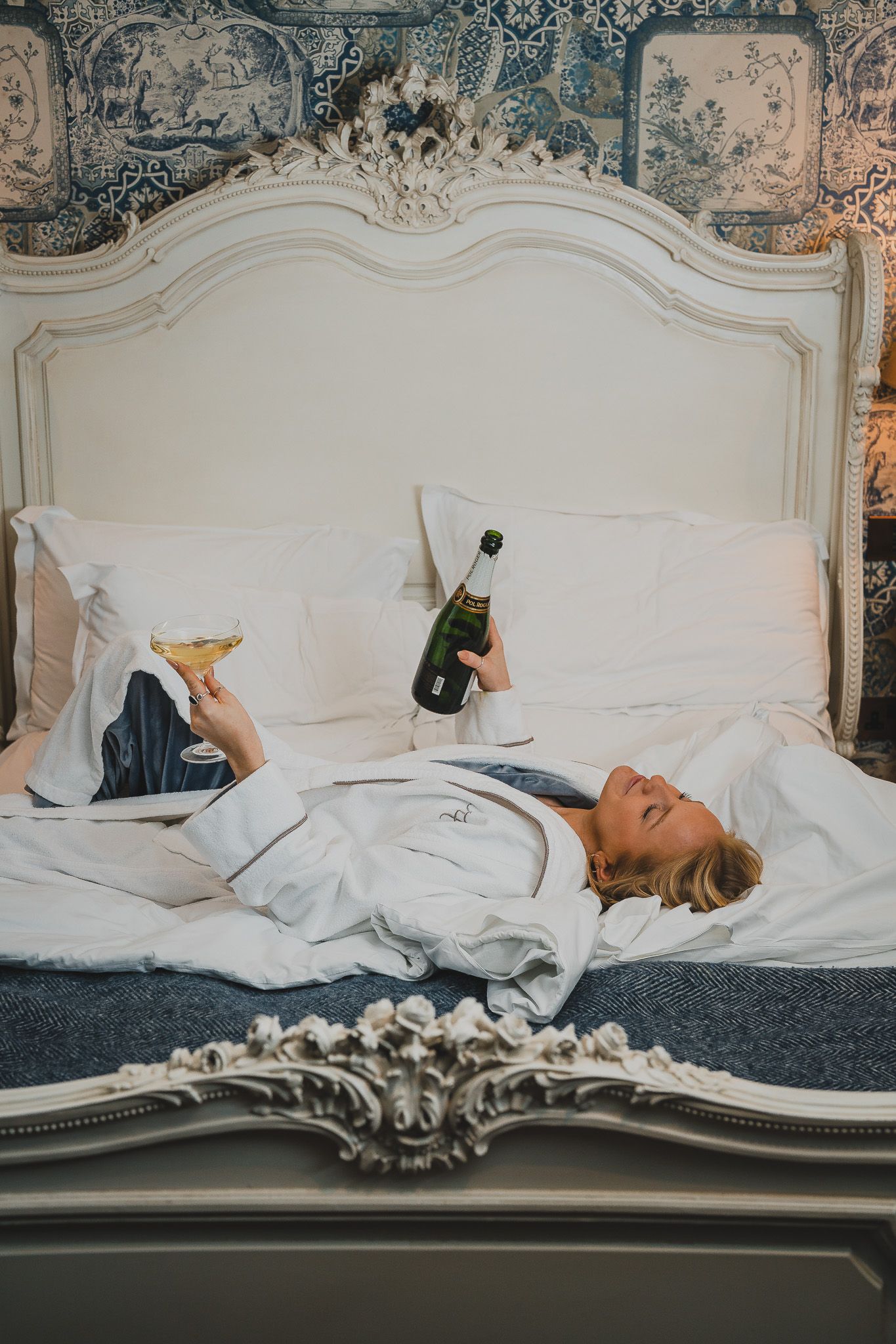 Woman in white robe relaxing on luxury hotel bed holding champagne bottle and glass.