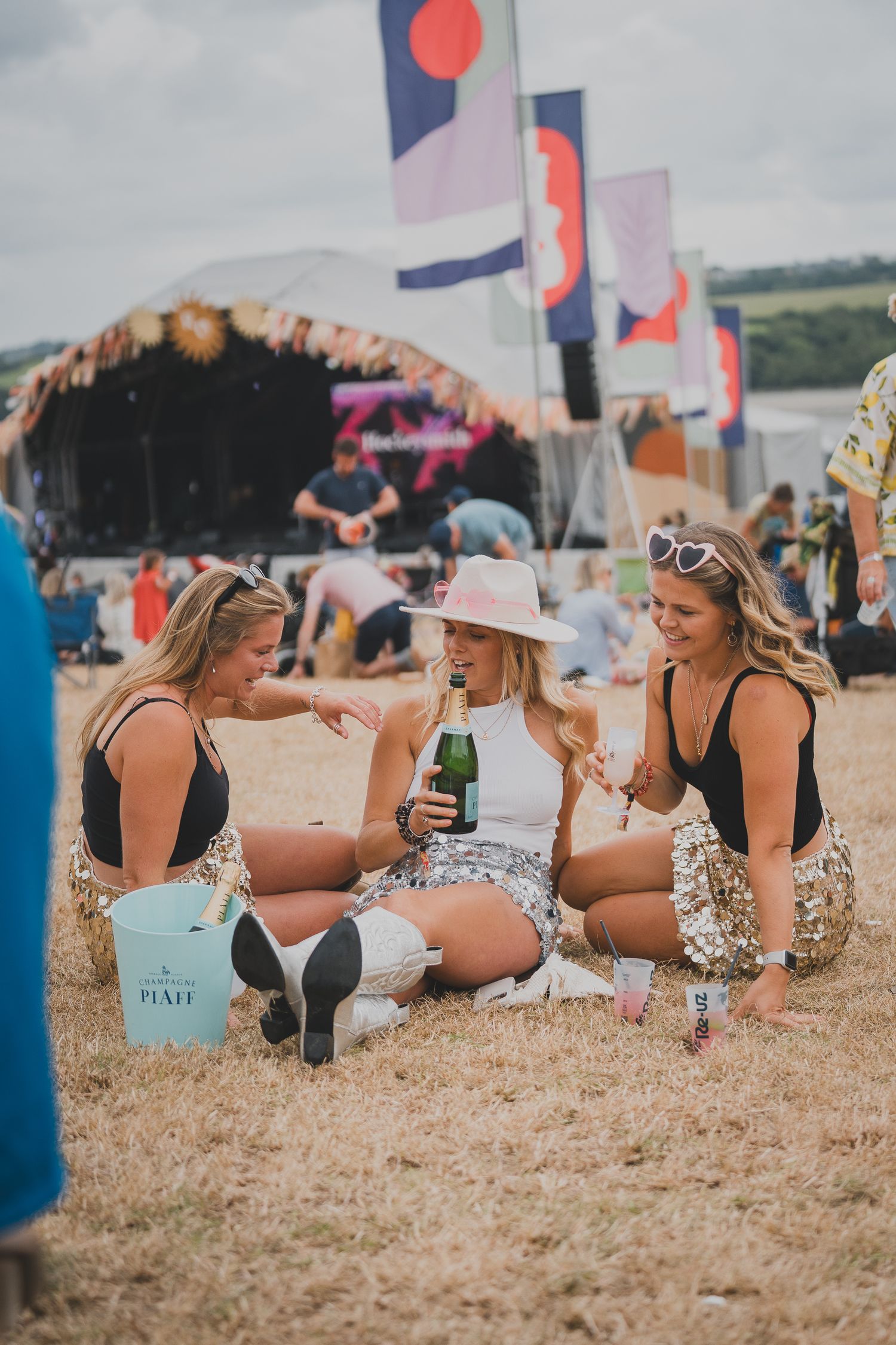 Three women sitting on grass at outdoor music festival, sharing champagne and celebrating in sequin outfits.