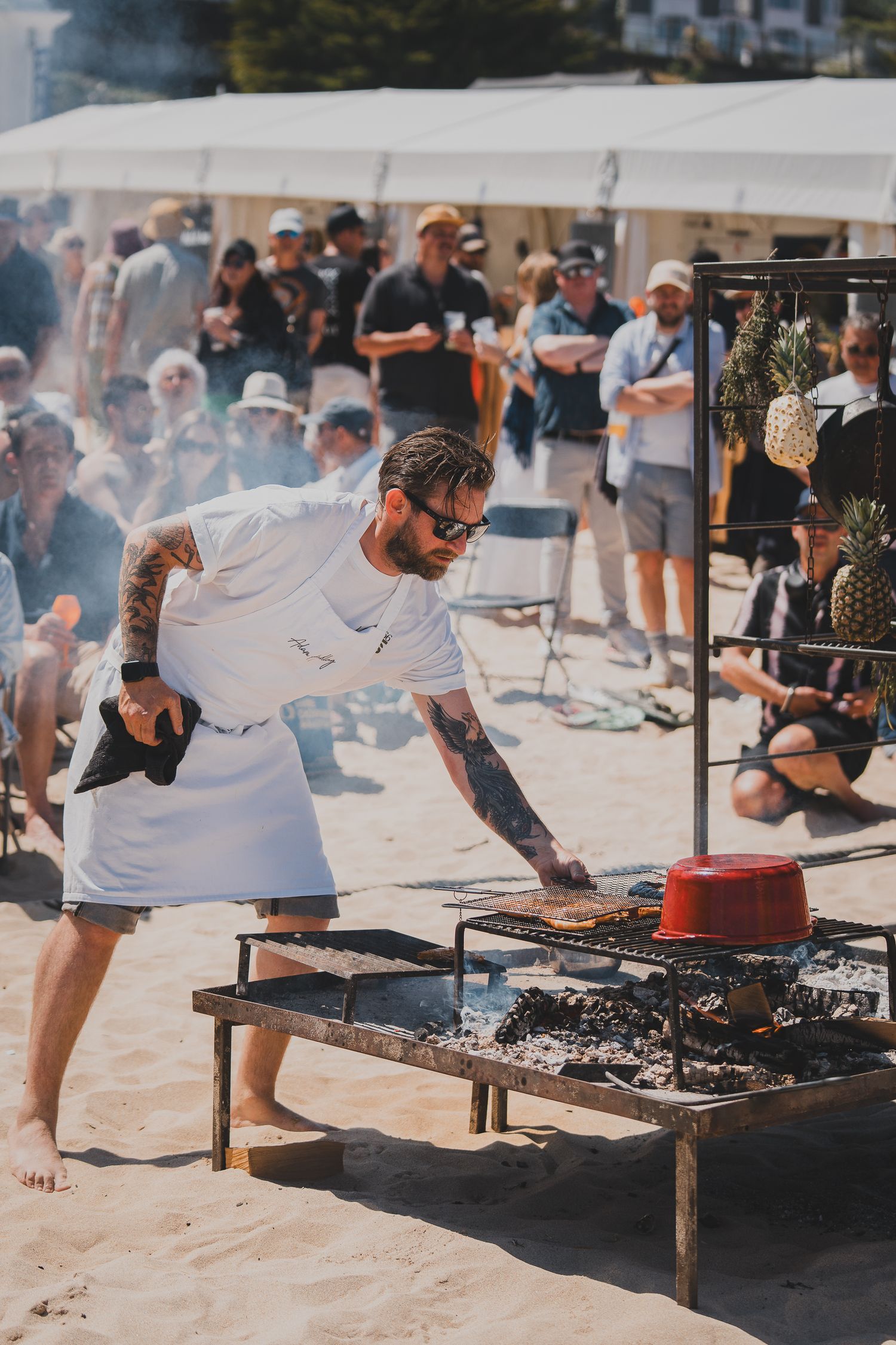 Tattooed chef in white apron grills food on a beach barbecue before a crowd of spectators.