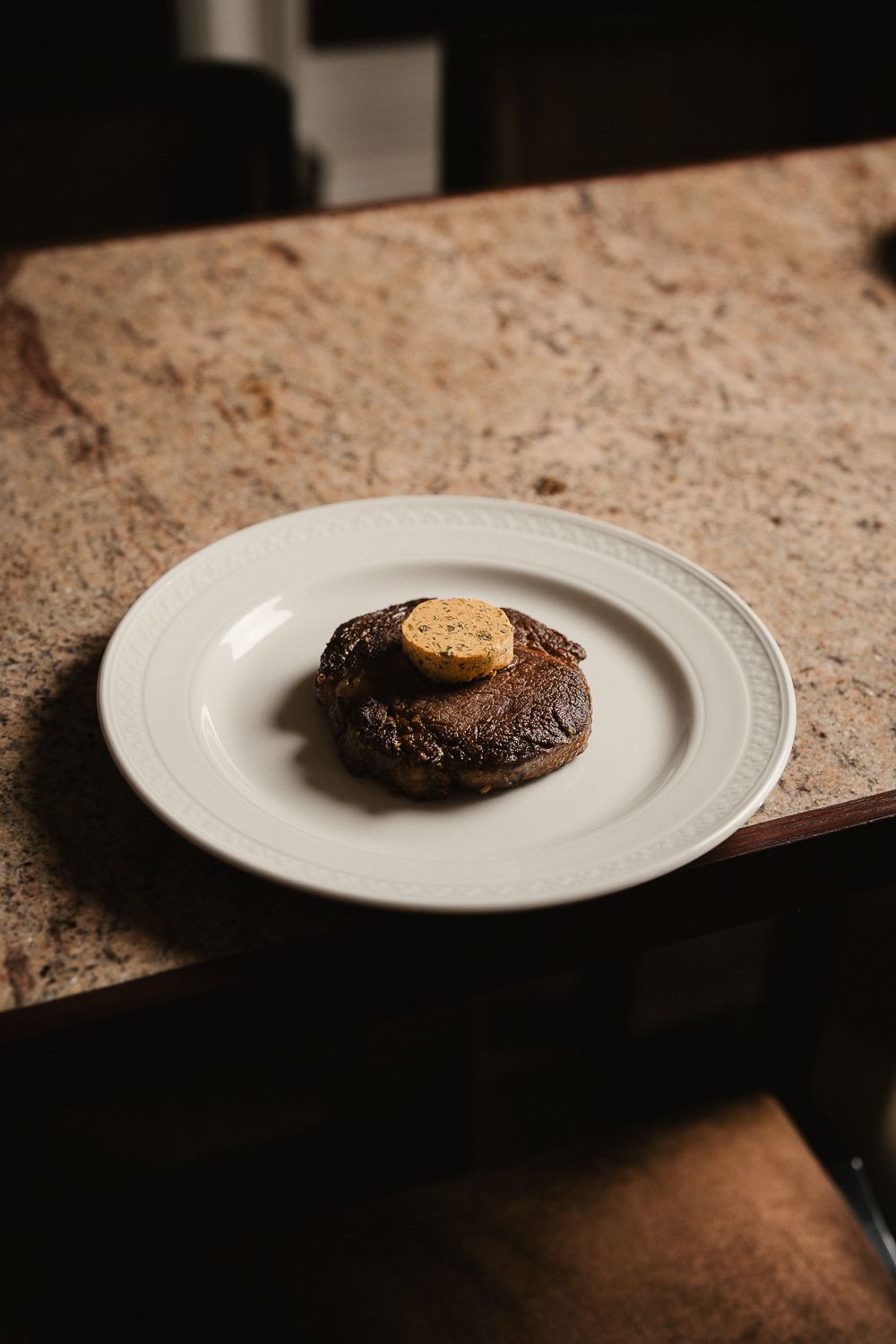 Perfectly seared steak medallion topped with herb butter on white plate on marble surface.