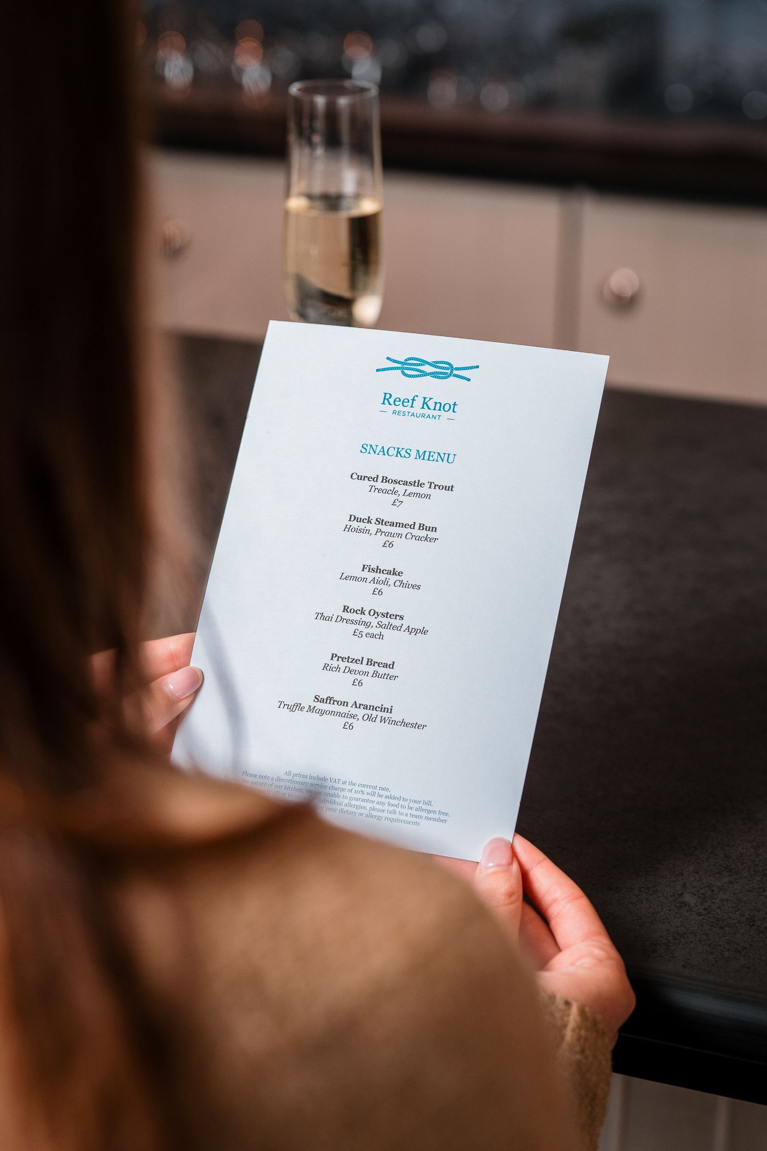 Person holding Reef Knot restaurant snacks menu with champagne glass visible in background.