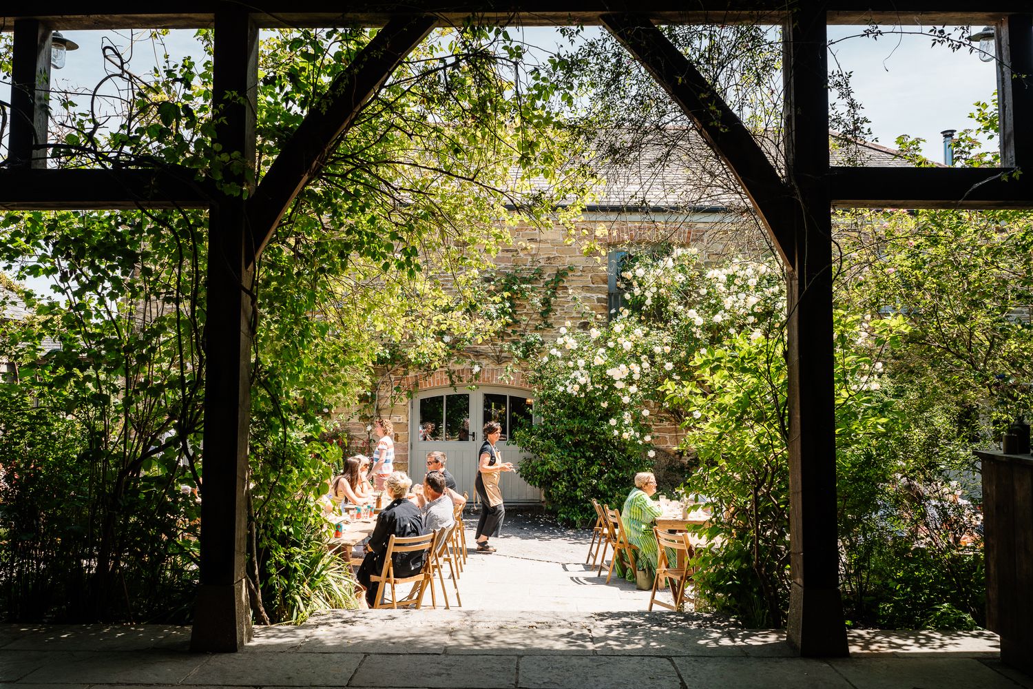 People dining outdoors in a lush garden courtyard framed by a wooden arch at a rustic stone building.
