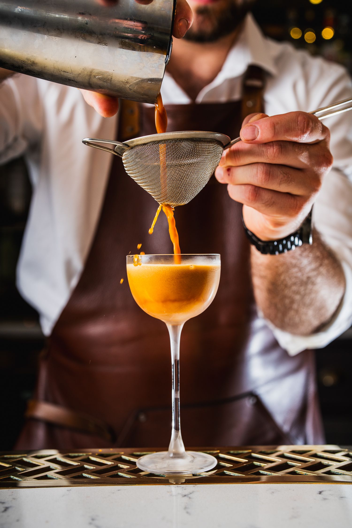 Bartender double straining an orange cocktail into a stemmed coupe glass.