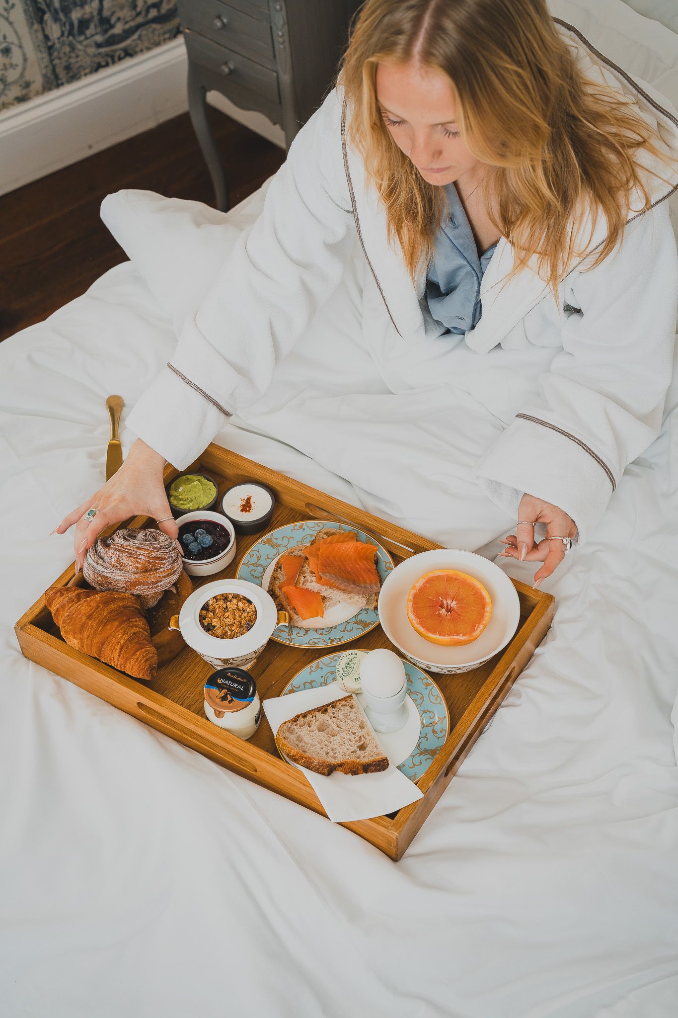 Woman in white bathrobe enjoying breakfast in bed with croissant, salmon, grapefruit, and coffee on wooden tray.