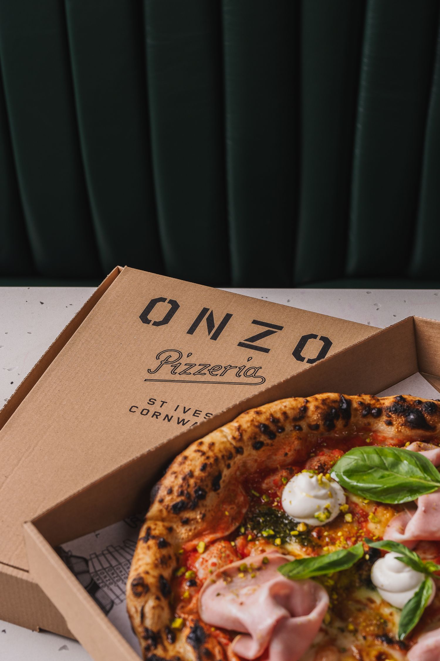 Neapolitan pizza with ham and basil resting on an Onzo Pizzeria branded takeaway box in St Ives Cornwall.
