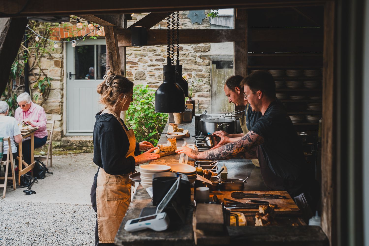 Restaurant staff preparing food at an outdoor open kitchen surrounded by greenery and stone walls.