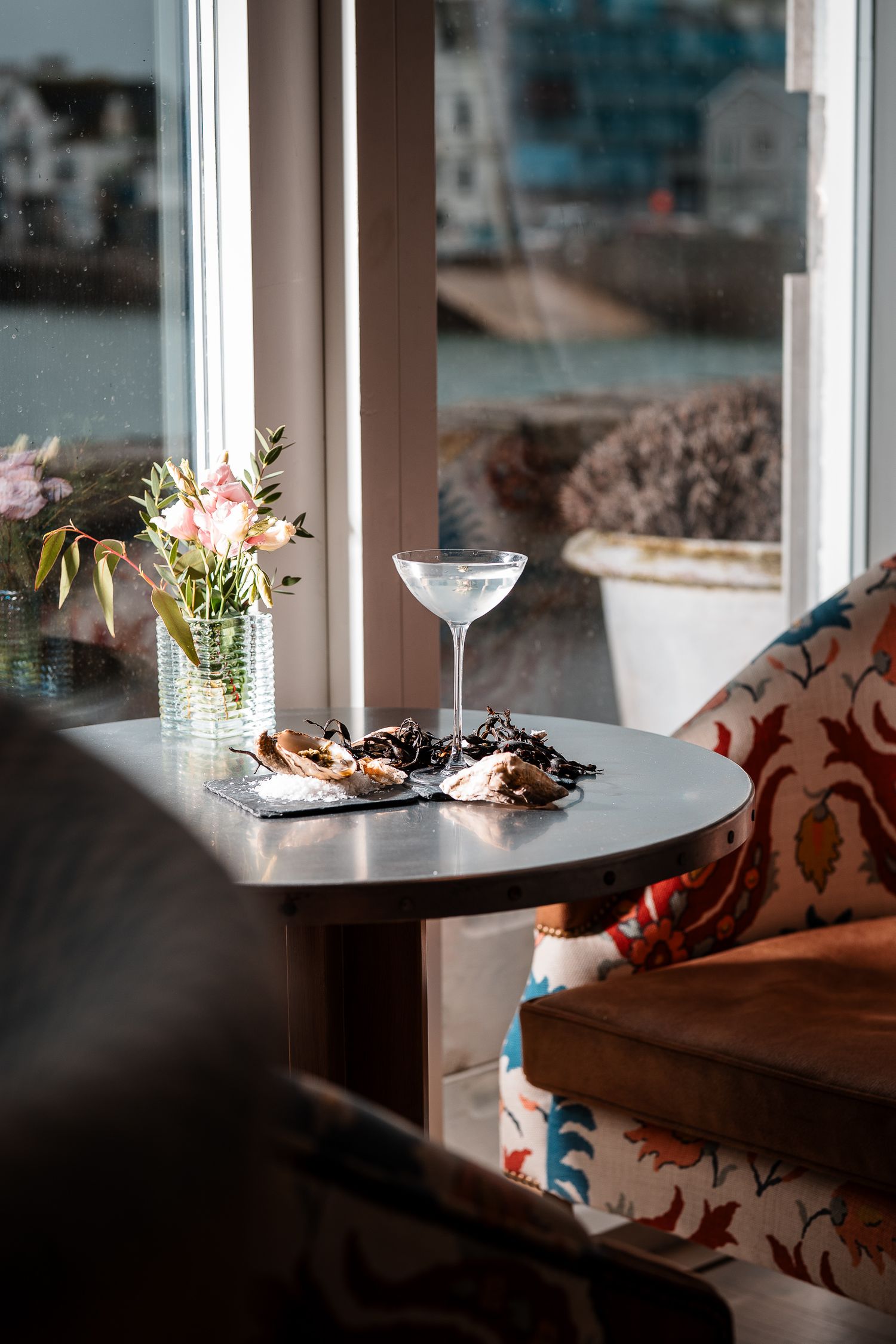 Cozy window seat with oysters, martini glass, and pink flowers overlooking coastal harbor view.