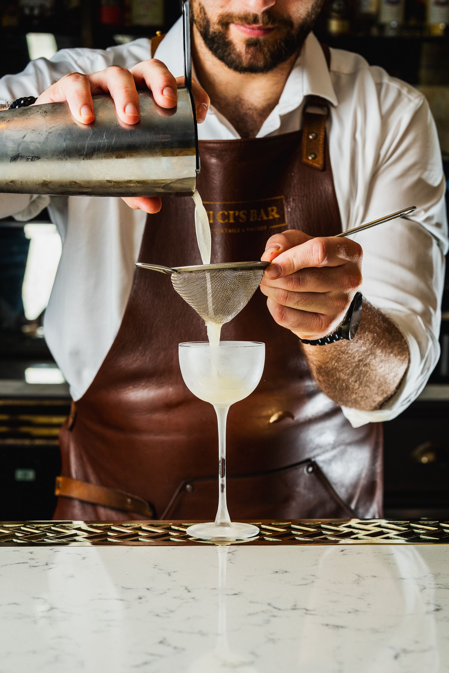 Bartender in leather apron straining a white cocktail into a coupe glass.