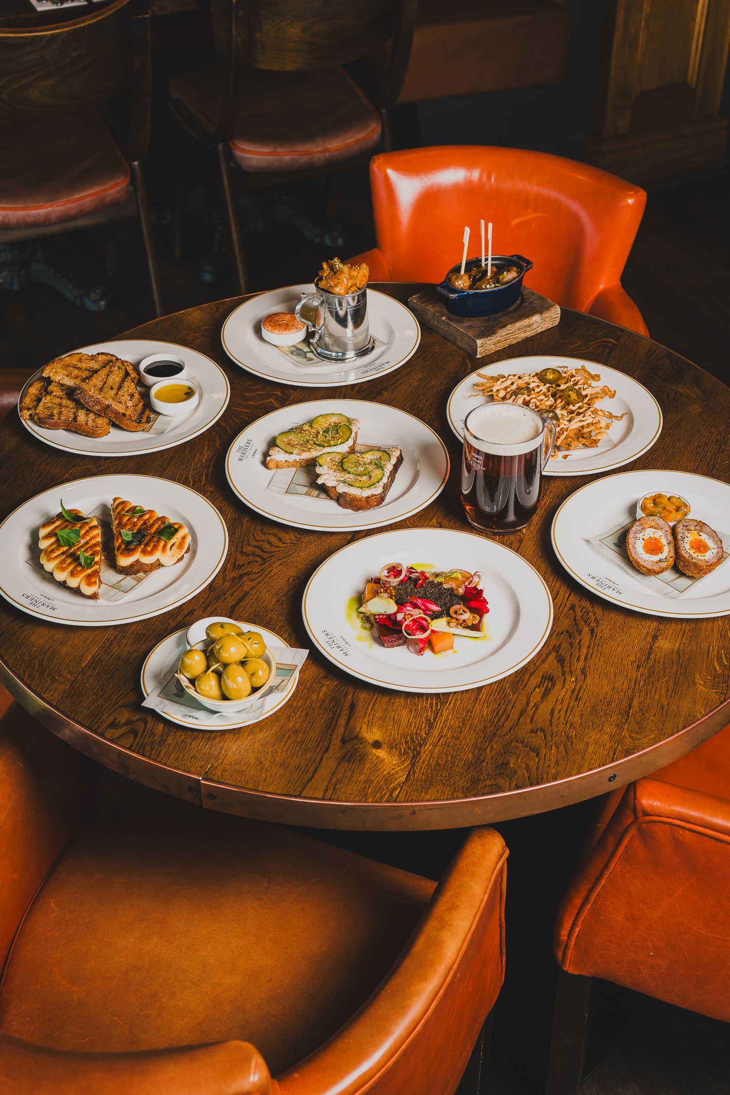 Variety of gourmet dishes and a pint of beer arranged on a round wooden restaurant table.