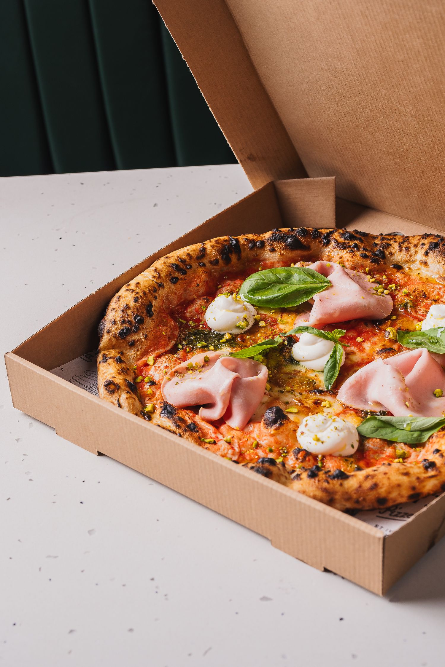 Freshly baked Neapolitan pizza with mortadella, mozzarella, and basil in an open cardboard takeaway box.