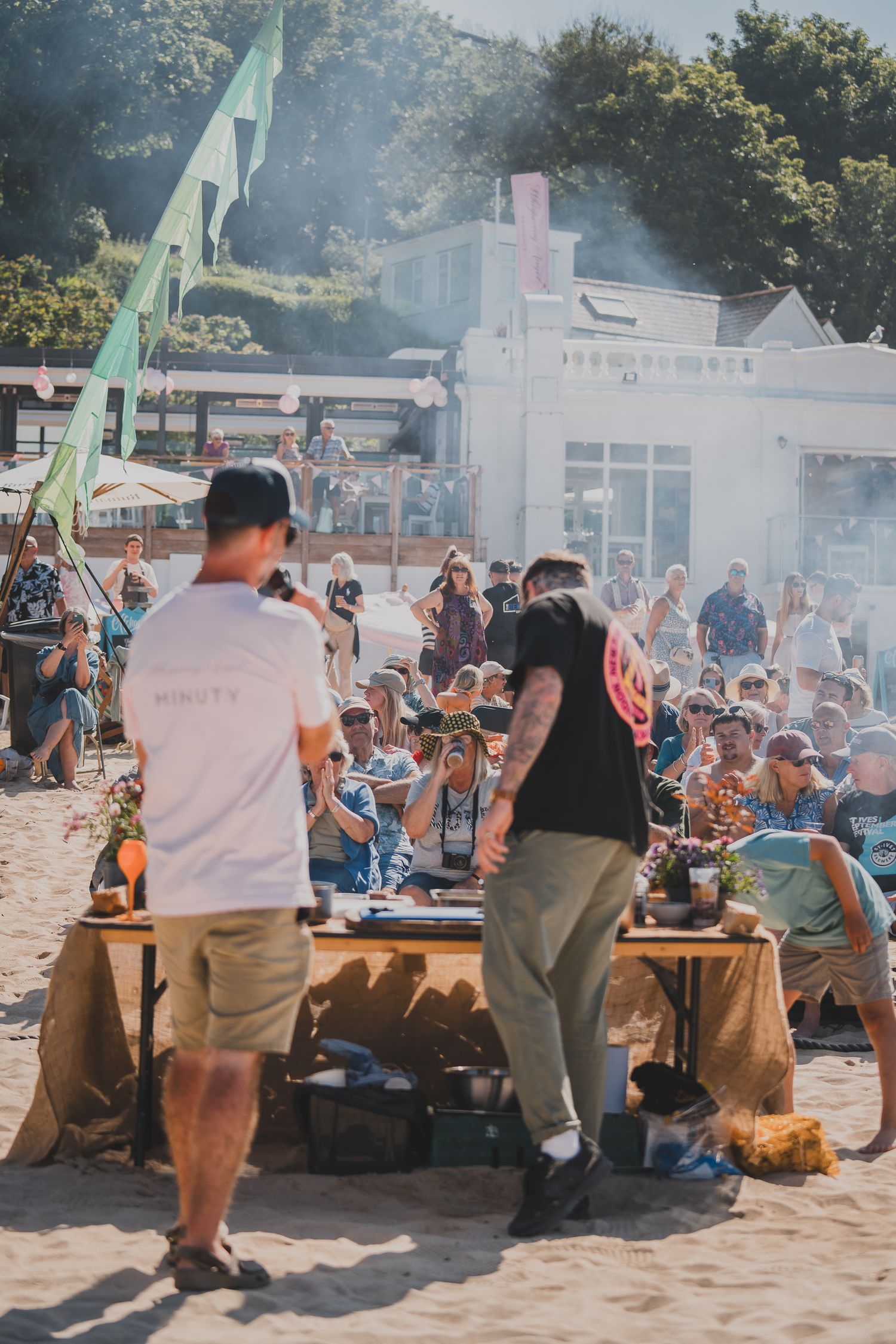 Two chefs cooking on beach for crowd at outdoor seaside food festival on sunny day.