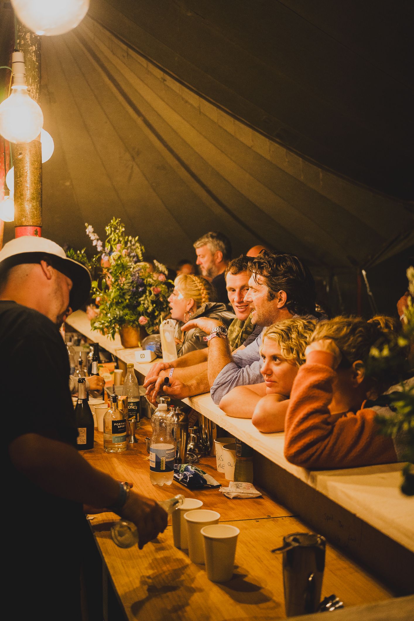 Festival guests ordering drinks at a warmly lit bar inside a canvas tent at a night event.