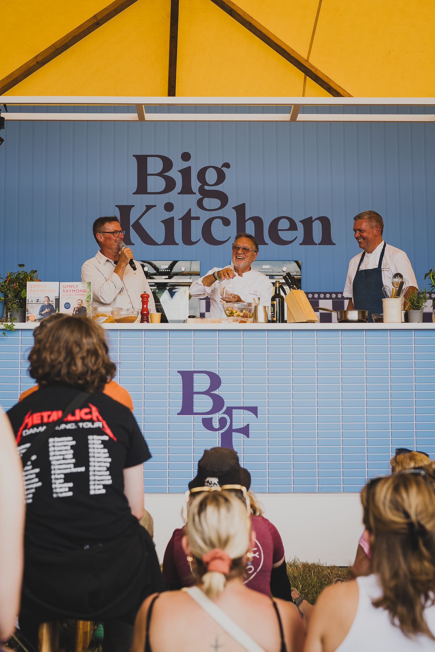 Three chefs presenting a live cooking demonstration at the Big Kitchen stage at a food festival.