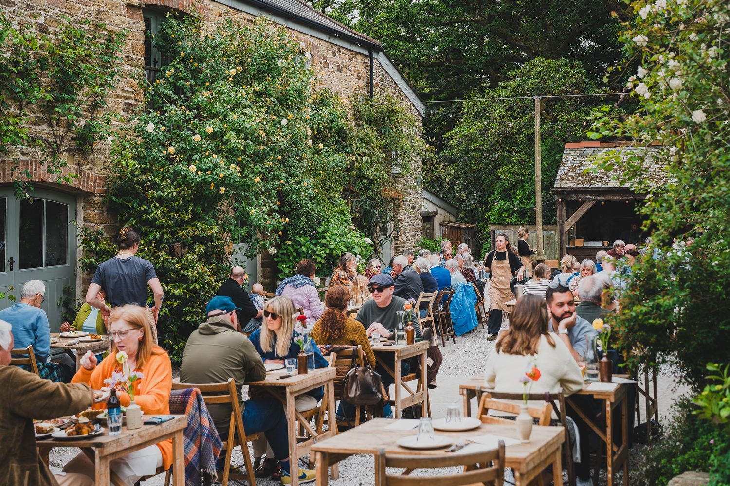Crowds of people dining outdoors at rustic stone courtyard restaurant surrounded by lush green foliage.