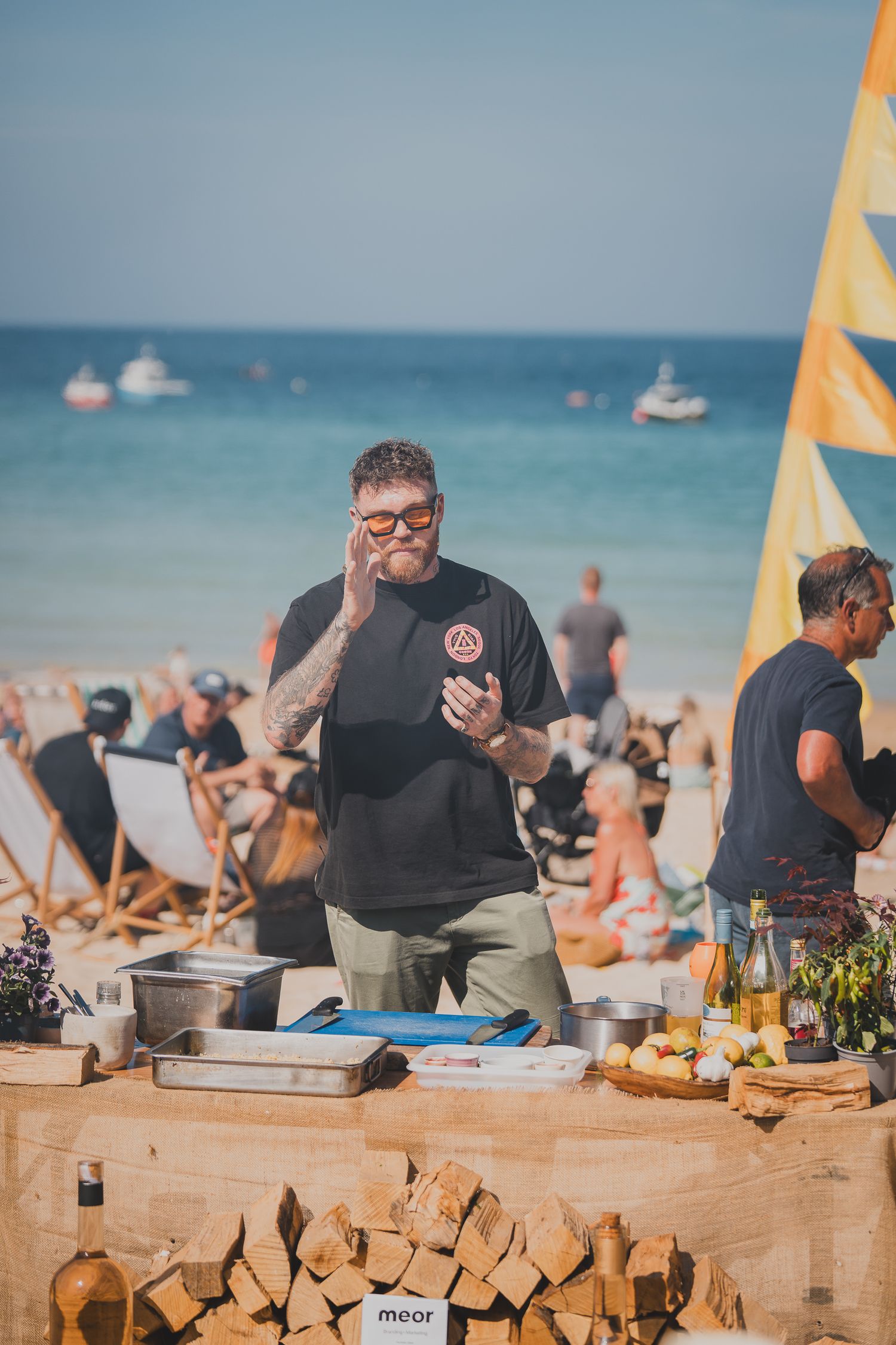 Tattooed chef presenting food at beachside cooking demonstration with ocean backdrop on sunny day.