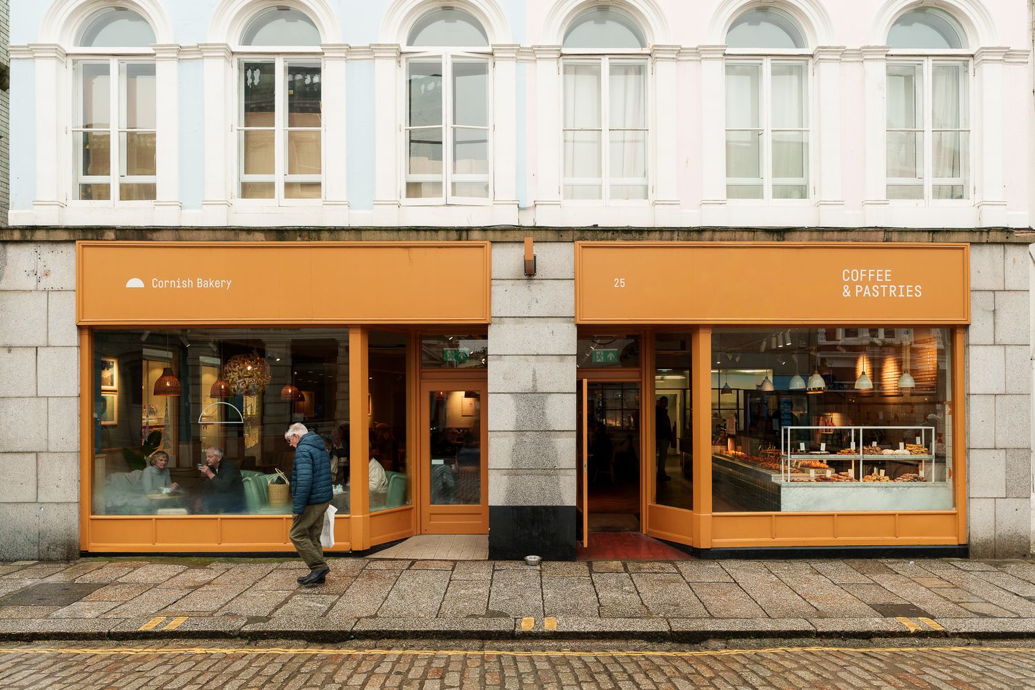 Wide shot of an orange Coffee & Pastries cafe storefront on a cobblestone street with a pedestrian passing.