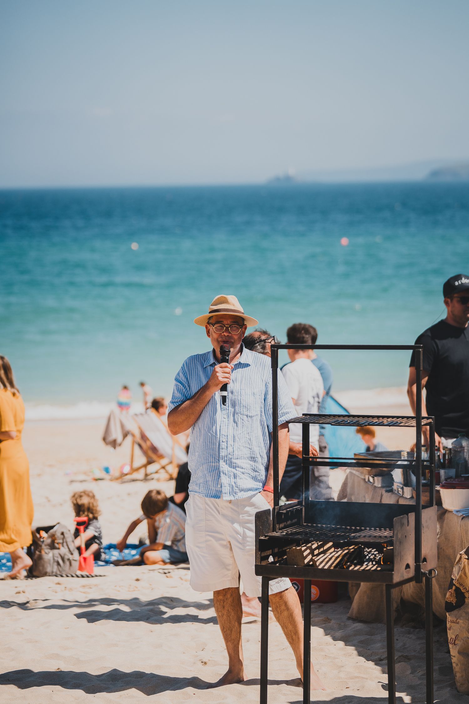 Man in straw hat speaks into microphone beside a beach grill with turquoise ocean in the background.