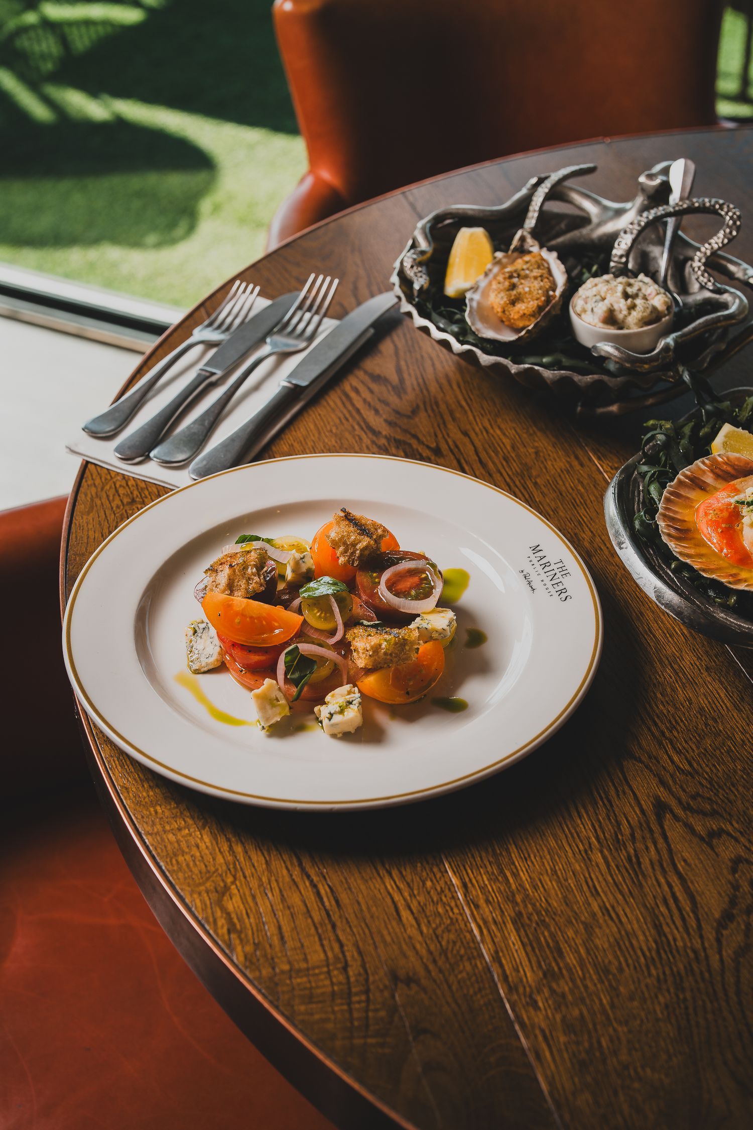 Fresh tomato and seafood salad on a branded plate beside oysters on a wooden restaurant table.