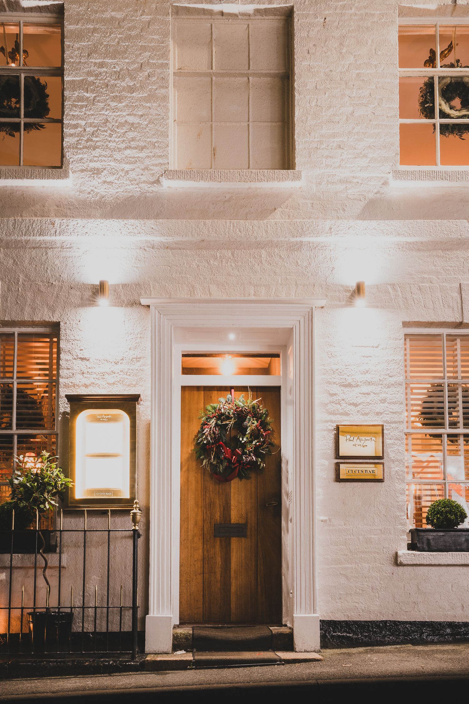 Charming white brick building entrance with wooden door, festive Christmas wreath, and warm wall lights at night.