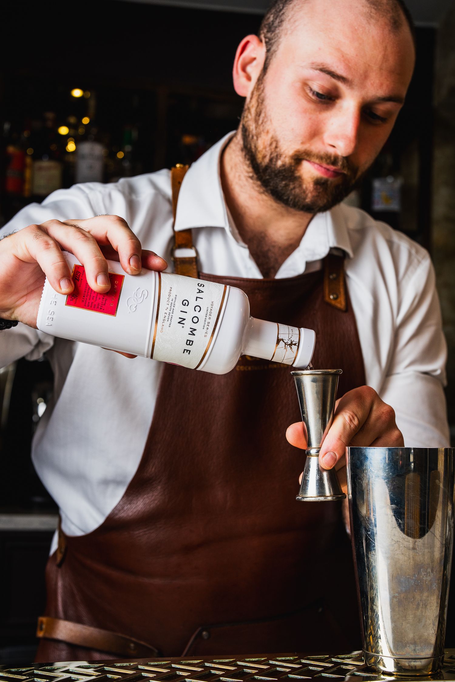Bearded bartender in leather apron pouring Salcombe Gin into jigger over cocktail shaker.
