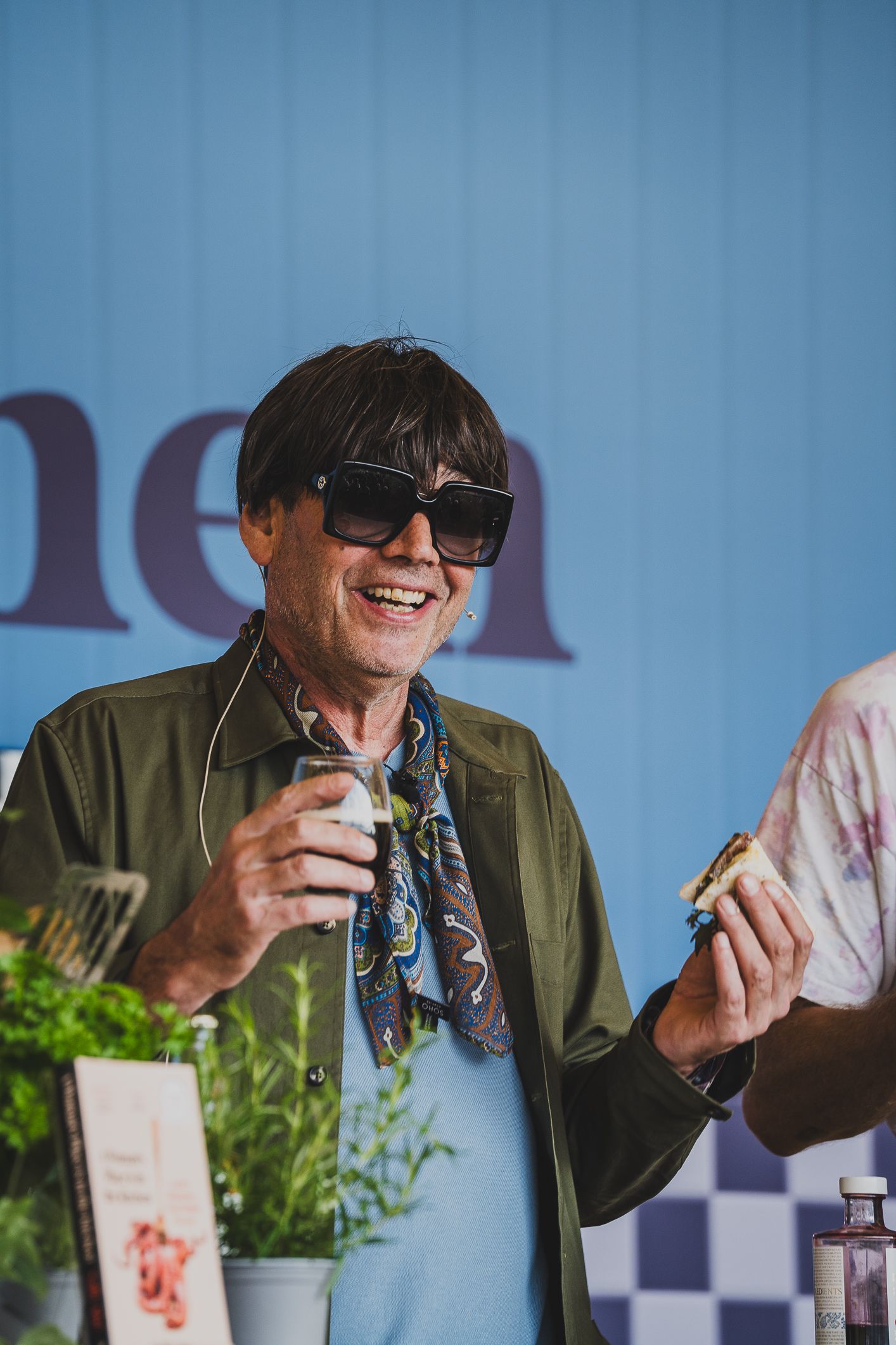 Smiling man wearing sunglasses and a green jacket enjoying food and drink at an outdoor festival.