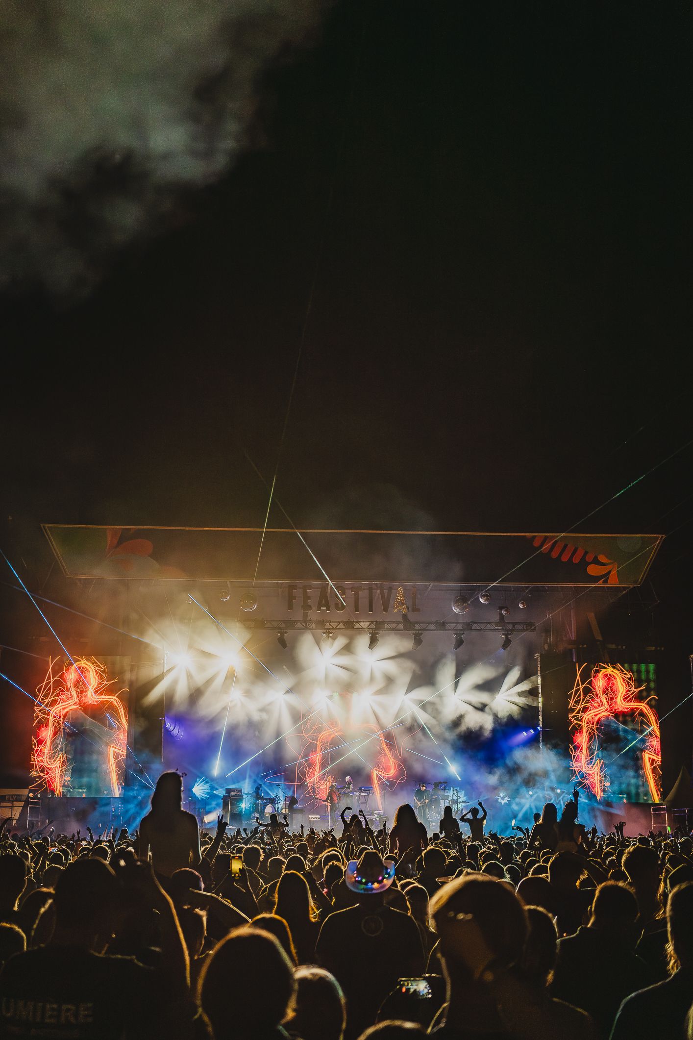 Massive outdoor festival crowd enjoying a spectacular light and pyrotechnic stage show at night.