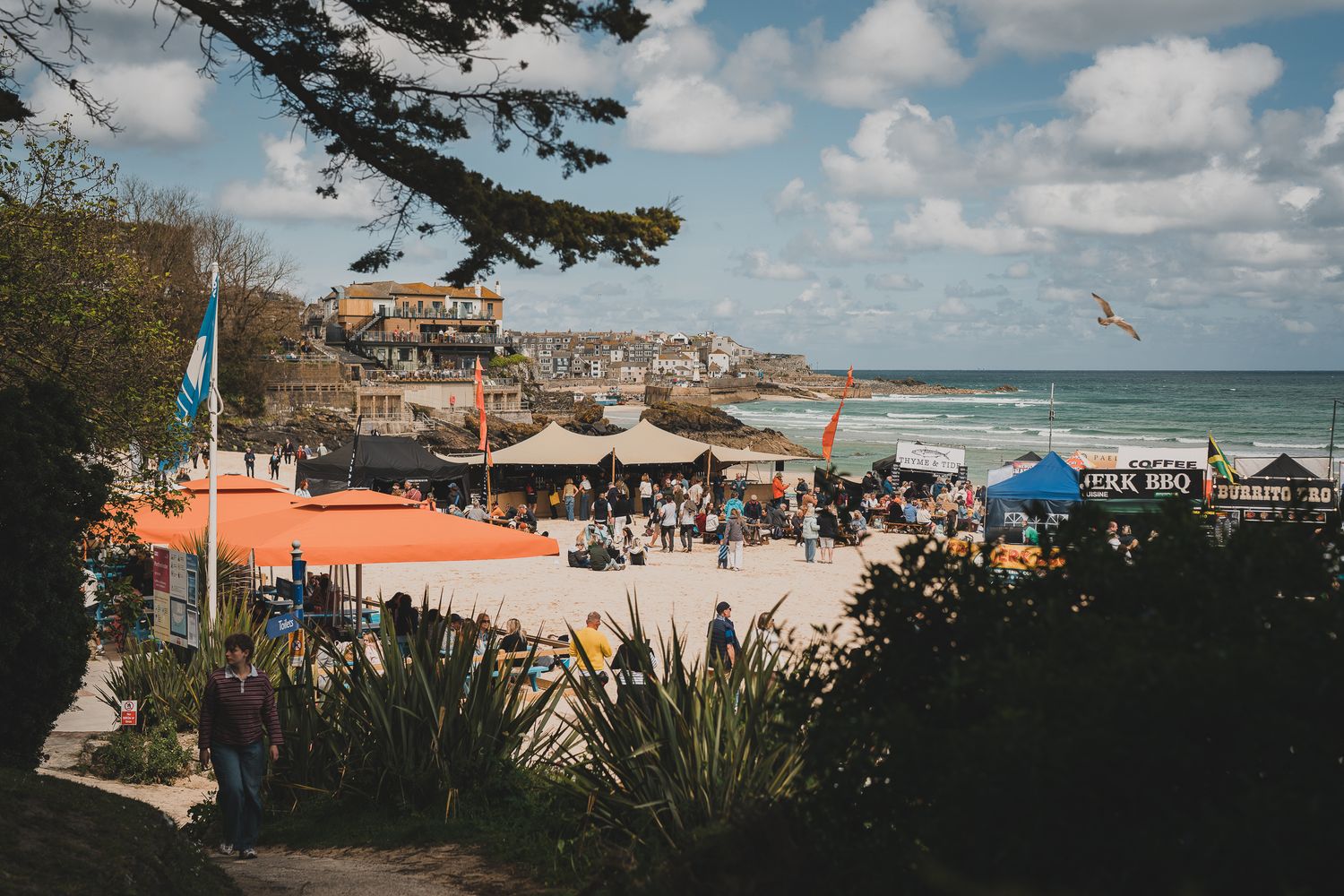 Aerial view of busy beach festival with colorful tents and stalls overlooking a stunning coastal town.