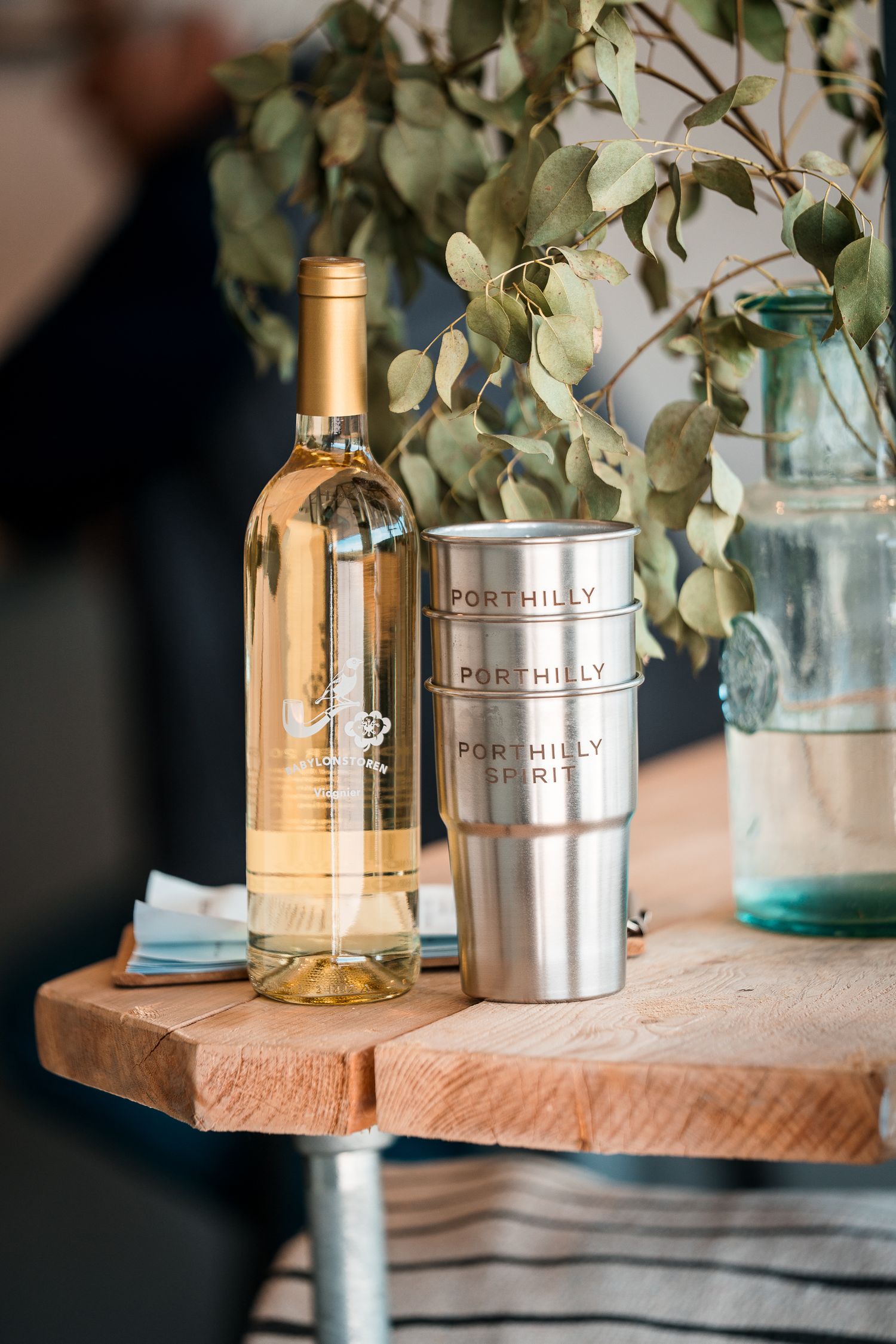 Porthilly Spirits stainless steel cups beside white wine bottle on rustic wooden table with eucalyptus decor.