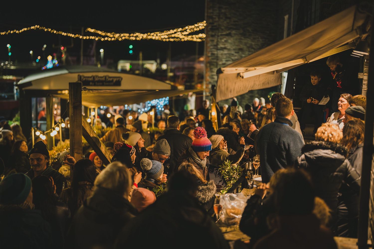 Large crowd enjoys a lively outdoor night market with string lights and food stalls.
