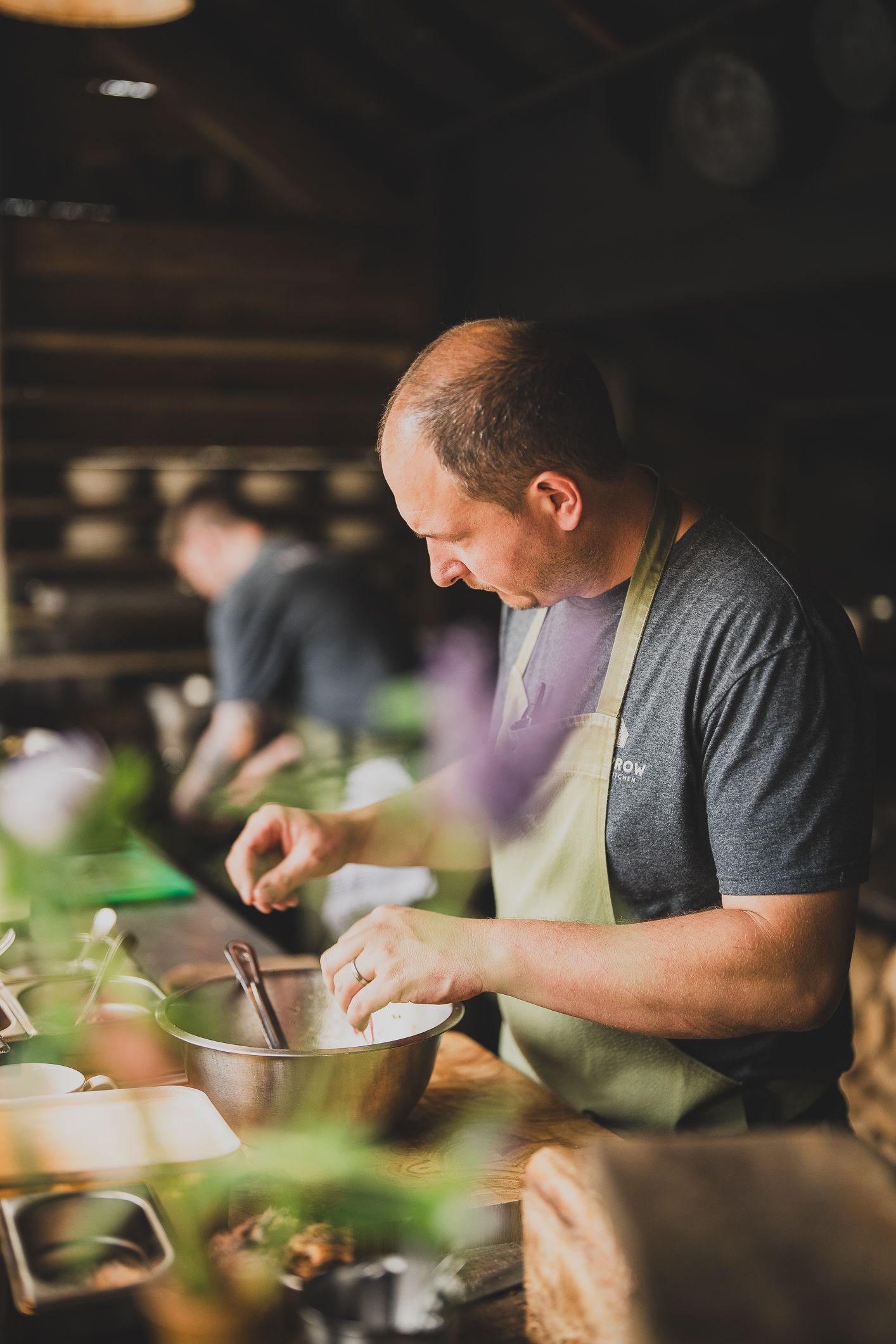 A chef in a green apron carefully prepares ingredients in a mixing bowl in a rustic kitchen.