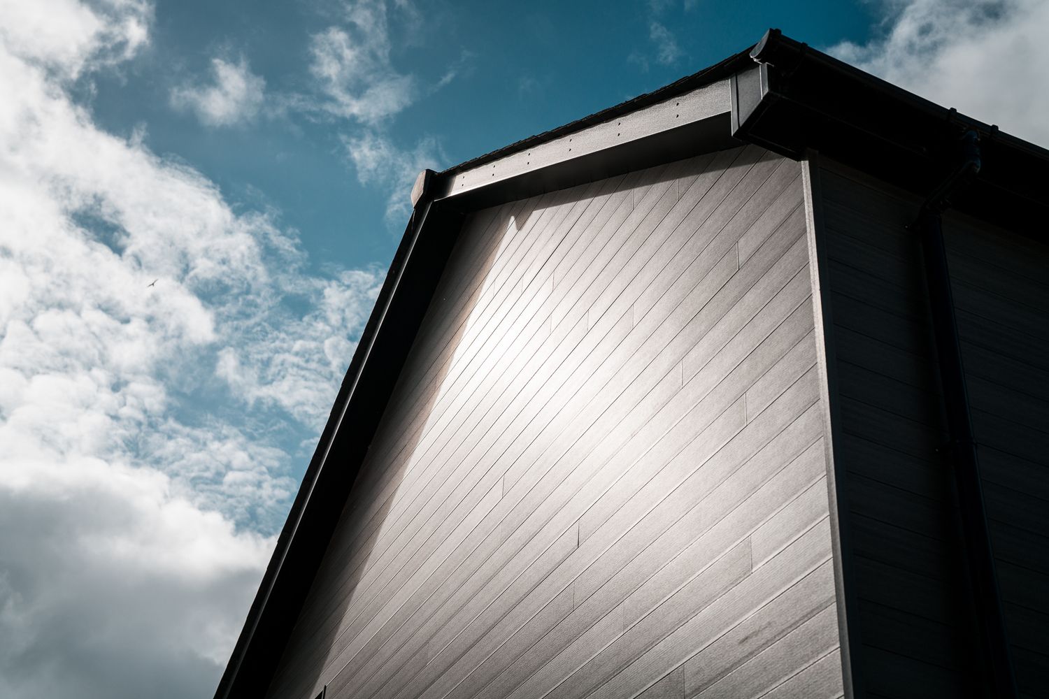 Low angle view of a modern building corner with metal cladding against a bright blue cloudy sky.