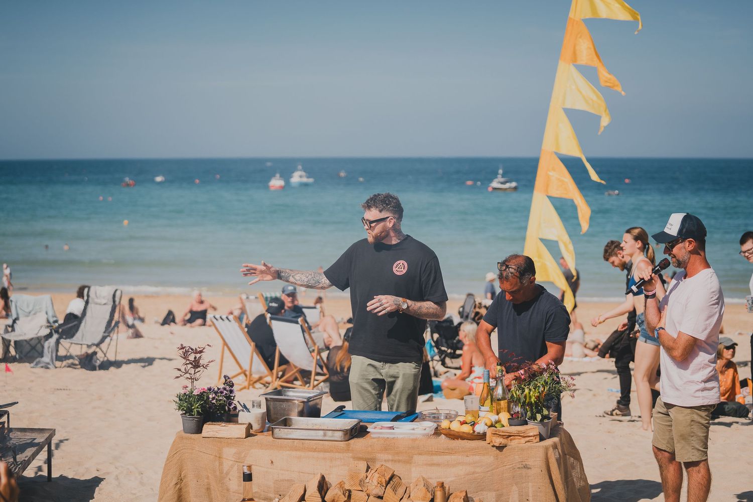 Chefs preparing food at outdoor beach cooking demonstration with yellow flag and sea in background.