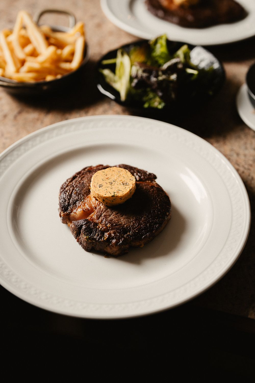 Grilled ribeye steak topped with herb butter served with fries and salad on white plate.