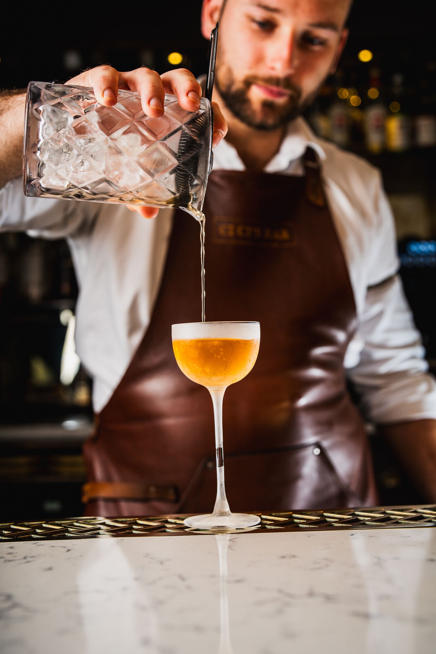 Bartender pouring an amber cocktail from a crystal mixing glass into a coupe.