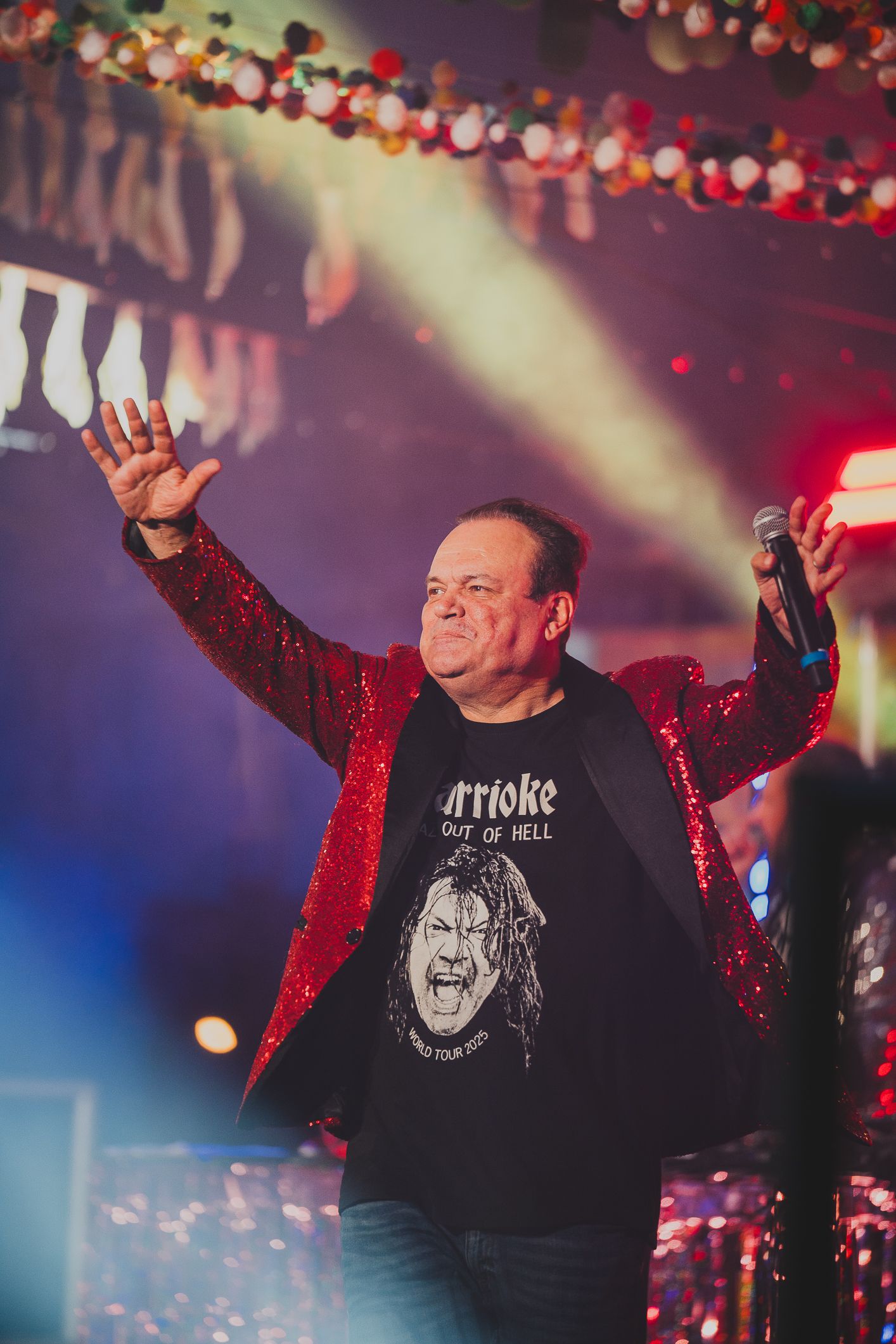 A performer in a red sequin jacket waves to the crowd while holding a microphone on a festive stage.