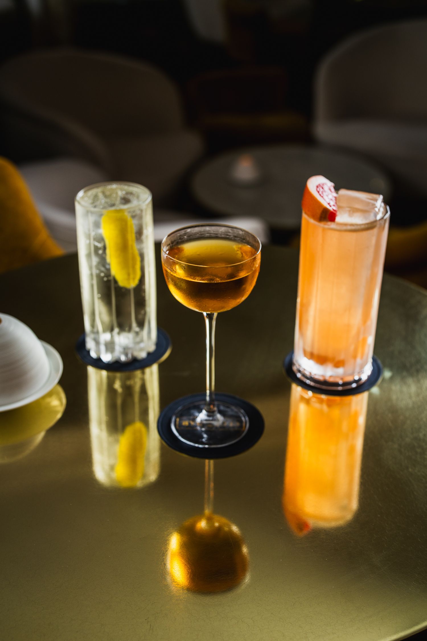 Three cocktails on gold reflective table including coupe and highball glasses in moody bar setting.
