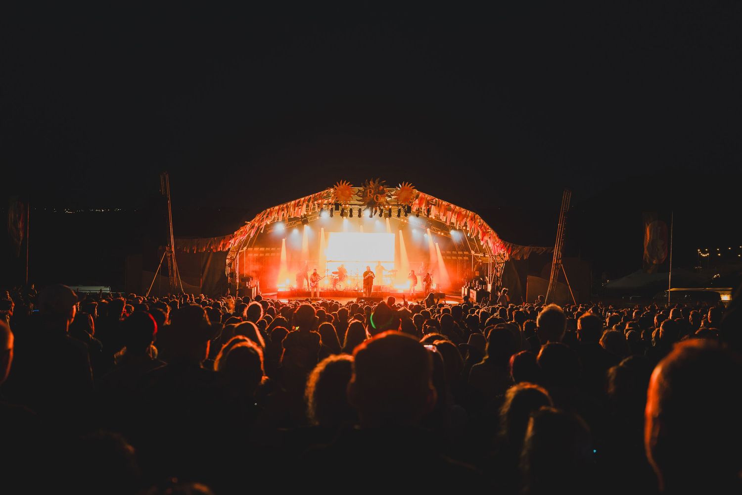 Large crowd faces a brightly lit outdoor festival stage glowing orange at night.