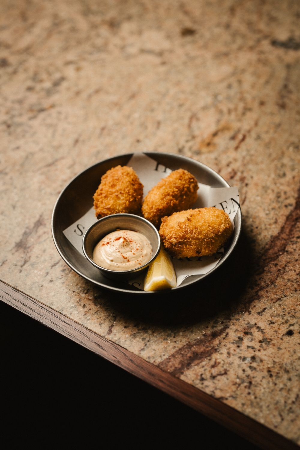 Golden fried croquetas served with spiced aioli dipping sauce and lemon wedge in metal bowl.