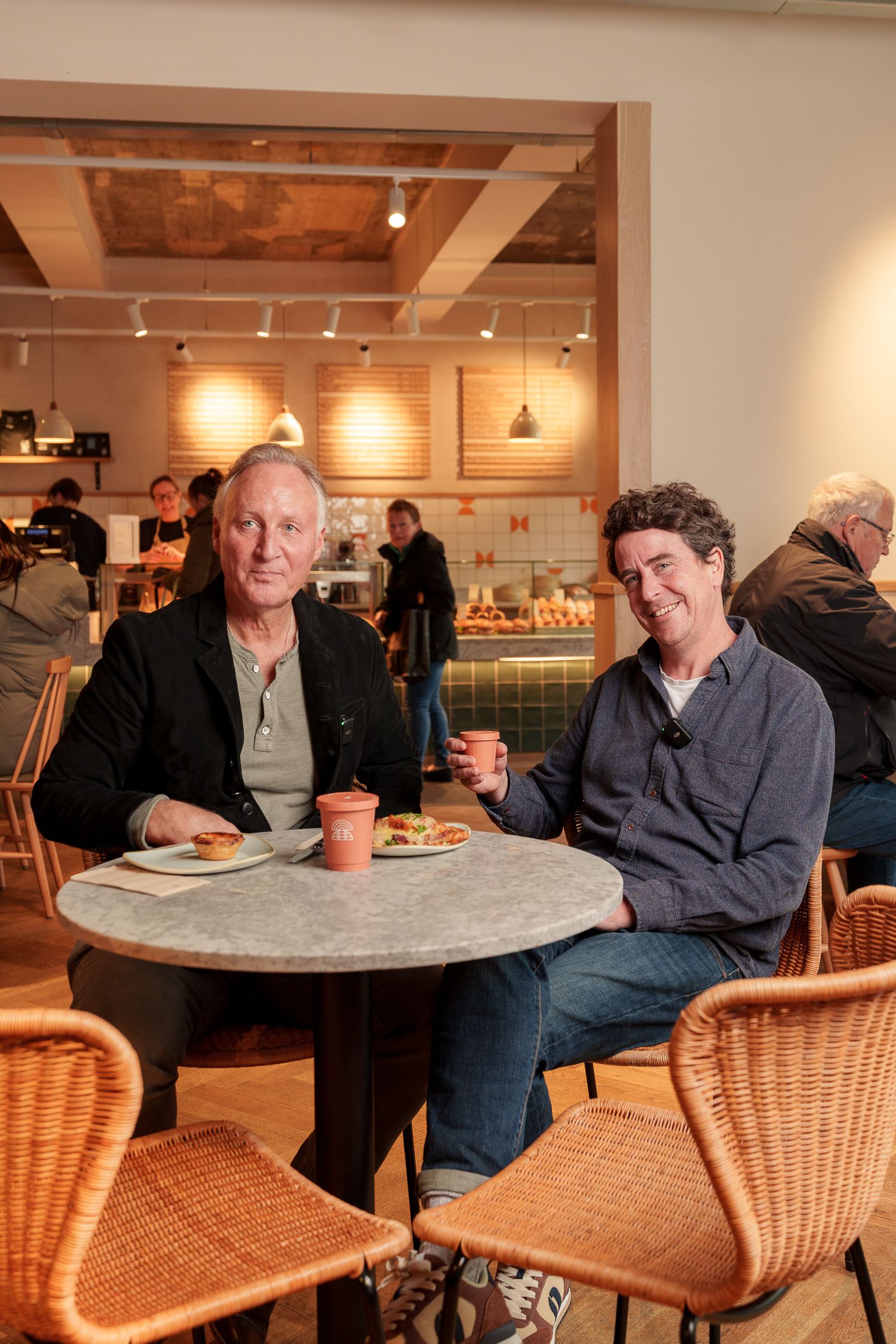 Two men smiling and enjoying coffee and food at a marble table in a warm bakery cafe.